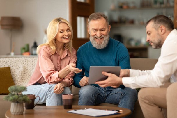 Professional Male Designer Showing Digital Tablet To Mature Clients Couple Offering Services Sitting On Sofa Indoor. Spouses Consulting With Real Estate Agent. Selective Focus - Valokuva, kuva