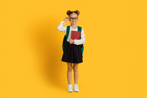 School Kid. Portrait Of Cute Little Girl With Backpack And Workbooks Posing Over Yellow Studio Background, Beautiful Smiling Preteen Schoolgirl Touching Eyeglasses And Looking At Camera, Copy Space - Фото, зображення