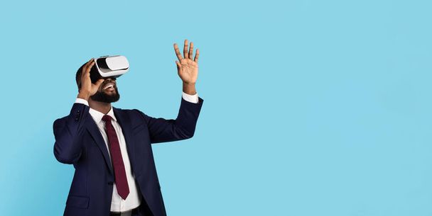 Virtual Reality Experience. Amazed Black Businessman speelt videospel in VR-brillen, opgewonden Afro-Amerikaanse man in pak staande over Blue Studio achtergrond, Panorama met kopieerruimte - Foto, afbeelding