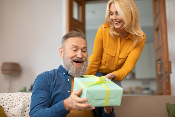 Volwassen vrouw geven geschenkdoos aan opgewonden echtgenoot feliciteren hem op verjaardag zitten op de bank, vieren familievakantie thuis. Cadeaus Aanbod Concept. Selectieve focus - Foto, afbeelding