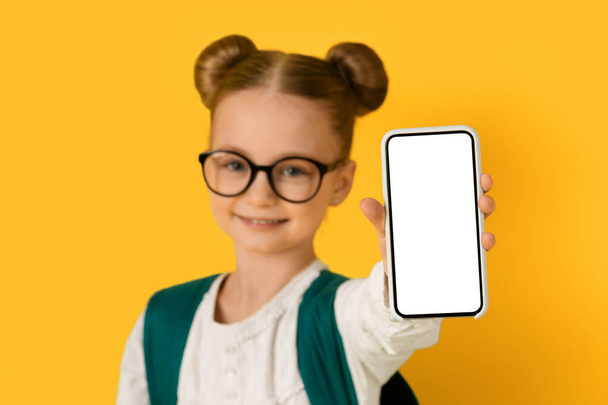 Cute Little Girl In Eyeglasses Showing Blank Smartphone With White Screen In Her Hand, Cheerful Preteen Female Child Recommending New Mobile Application While Standing Over Yellow Background, Mockup - Фото, изображение