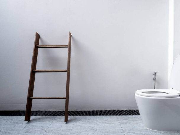 White restroom interior design, minimal style. Wooden towel rack, dark brown standing towel ladder near ceramic flush toilet and bidet shower on square tiles and white wall background with copy space. - Foto, Imagem