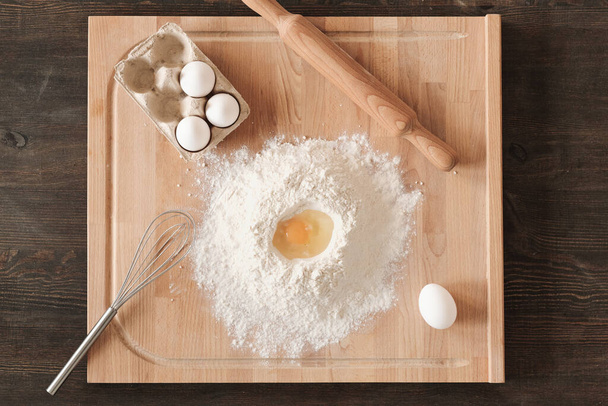 Directly above view of wooden board with flour, eggs, whisk and rolling pin on kitchen counter - Fotó, kép