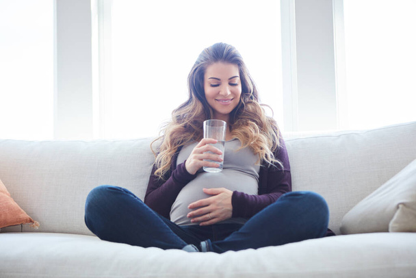 Varmistan, että vauva on nesteytetty. Täyspitkä laukaus houkutteleva nuori raskaana oleva nainen juomaveden istuessaan sohvalla kotona - Valokuva, kuva