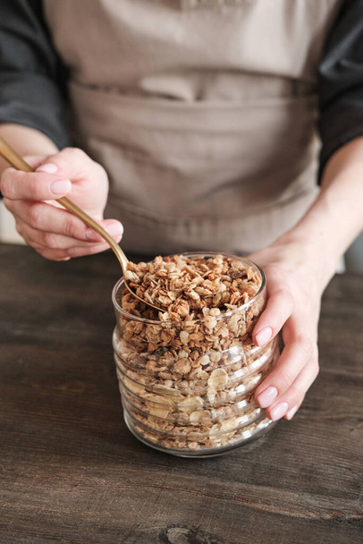Close-up of unrecognizable woman in apron standing at wooden table and preparing granola with muesli - Foto, Imagen