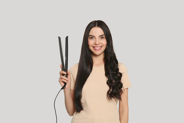 Beautiful happy woman with hair iron on light grey background - Photo, Image