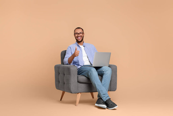 Usmívající se mladý muž s notebookem sedí v křesle a ukazuje palce nahoru na béžové pozadí - Fotografie, Obrázek