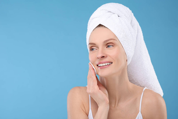 Beautiful woman in terry towel removing makeup with cotton pad on light blue background, space for text - Zdjęcie, obraz