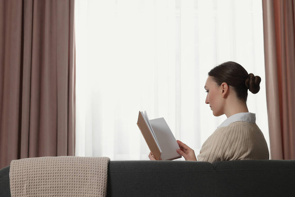 Woman reading book on sofa near window with stylish curtains at home. Space for text - Fotó, kép