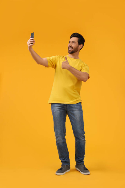 Smiling man taking selfie with smartphone and showing thumbs up on yellow background, space for text - 写真・画像