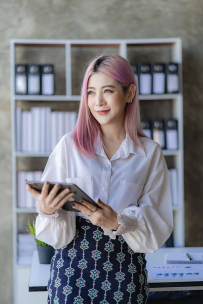 Attractive pink-haired Asian female employee standing in front of a desk Using a touch tablet computer, smiling, emailing business information to customers at a desk in the company's office. - Photo, Image