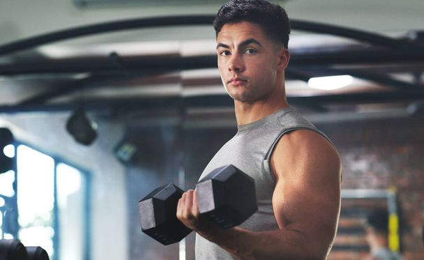 Aumentando de volumen y construyendo músculo. Retrato de un joven deportivo haciendo ejercicio con una mancuerna en el gimnasio - Foto, Imagen