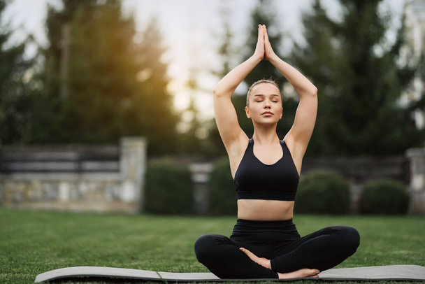 Spor kıyafetli genç bir kadın parkta yoga yapıyor. Sağlıklı bir kadın sabah lotus pozuyla çimlerde ve meditasyonda oturuyor. Sağlık, fitness, egzersiz ve iş hayatı dengesi kavramları. - Fotoğraf, Görsel