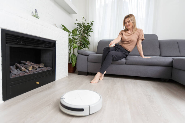 Modern robot vacuum cleaner near sofa with resting woman in room. - Photo, Image