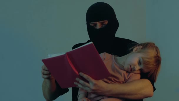 Male kidnapper while reading book for small girl hostage. Little girl sleeping in the hands of the criminal. Copy space. Child abduction concept. Slow motion - Footage, Video