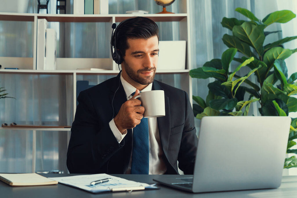Manager of call center operator office sitting on his desk with his coffee while working on laptop. Office worker wearing headset and black suit working on customer support or telemarketing. fervent - Φωτογραφία, εικόνα