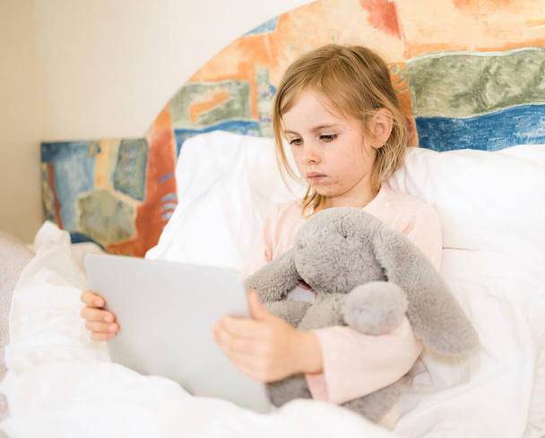 Sick child with red pimples sitting in bed hugging fluffy toy. Little girl looking at tablet. Chickenpox, varicella virus or vesicular rash on child's body and face - Foto, Bild