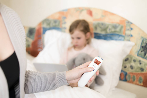 mother measuring temperature of her daughter. child got high temperature. Kid sitting in bed with tablet and furry toy and no face woman holding infrared thermometer which indicates 38,2 C - Photo, Image