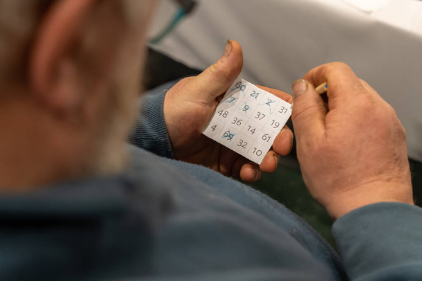 Por encima de la vista del hombro del anciano sosteniendo la tarjeta de bingo con números tachados y lápiz - Foto, imagen