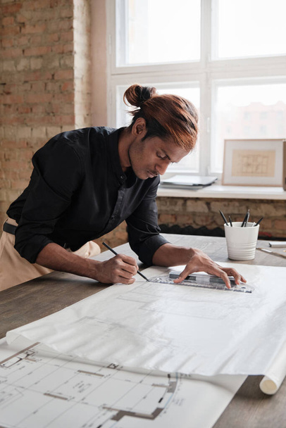 Focused young mixed race engineer with ponytail leaning on desk and using ruler while drawing house plan in office - Foto, afbeelding