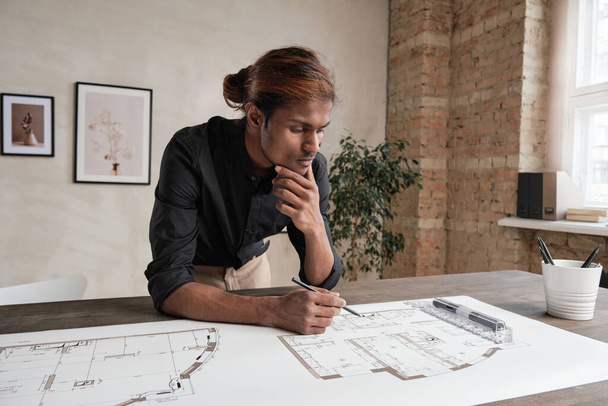 Thoughtful young mixed race architect with dyed hair leaning on table and analyzing sketch while thinking about design - Photo, image