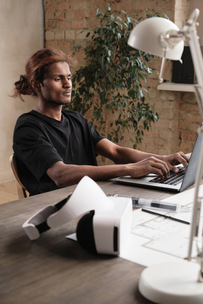 Content young Indian architect with ponytail sitting at wooden desk with virtual reality headset and developing design on laptop in loft office - Photo, image