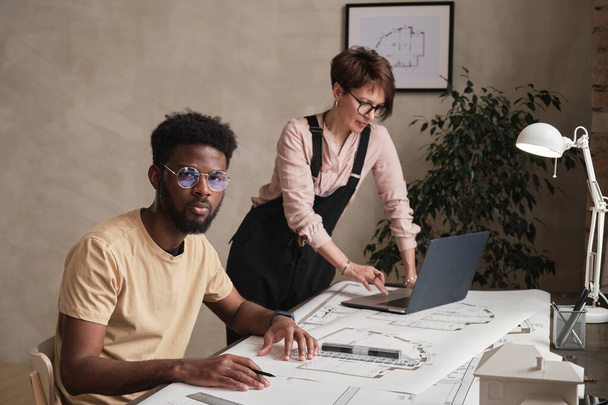 Portrait of serious young African-American man in tshirt sitting at table and working under control of experienced architect - Fotografie, Obrázek