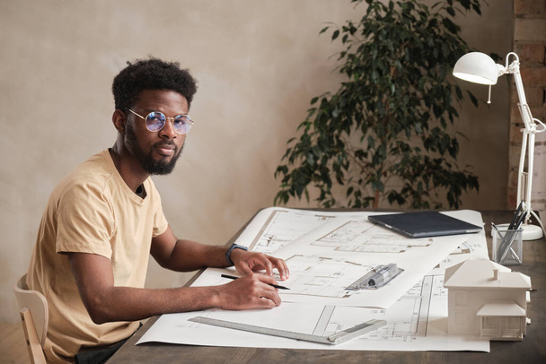 Portrait of content creative African-American architect in round-shaped glasses sitting at table with blueprints in office - Foto, imagen