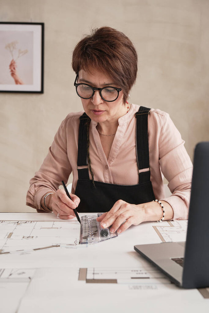 Concentrated mature woman in glasses sitting at table and using ruler while making floor plan in office - Φωτογραφία, εικόνα