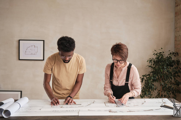 Concentrated Interracial design architects in eyeglasses standing at table in office and examining building plan together - Fotó, kép