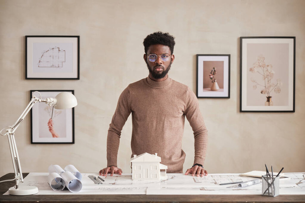 Portrait of serious confident African American architect in round-shaped eyeglasses standing at table with blueprints in office - Fotoğraf, Görsel