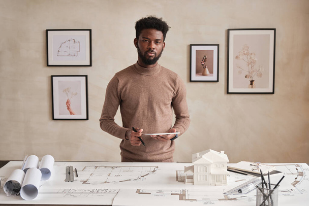 Portrait of serious young bearded black architect in sweater standing with tablet at table full of sketches and supplies in contemporary office - Foto, imagen