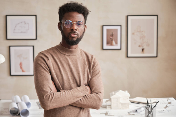 Portrait of serious young African American architect in round-shaped eyeglasses standing with crossed arms in modern office with pictures on wall - Valokuva, kuva