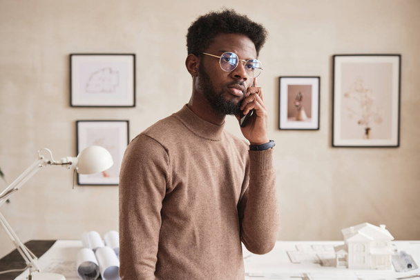 Serious young African American building architect in round-shaped glasses standing against table with D model of house and talking by phone - Photo, image