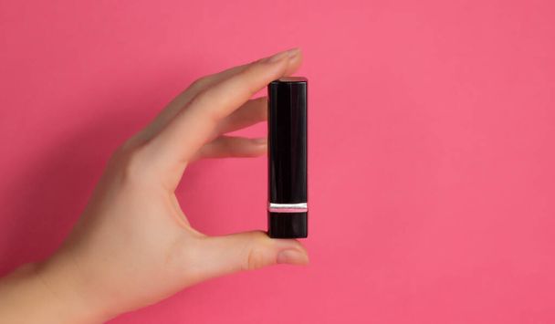 A womans hand shows a black tube of closed lipstick on a pink background - Fotó, kép