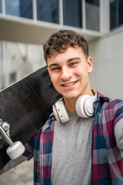One man young adult caucasian teenager stand outdoor with skateboard on his shoulder and headphones posing portrait looking to the camera happy confident wear shirt casual real person copy space - Photo, Image