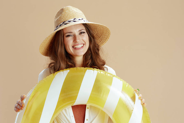 Beach vacation. smiling elegant middle aged housewife in white blouse and shorts isolated on beige background with inflatable circle and summer hat. - Valokuva, kuva