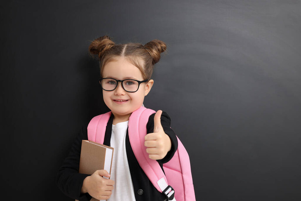 Happy little school child with notebook showing thumbs up near chalkboard. Space for text - Foto, afbeelding