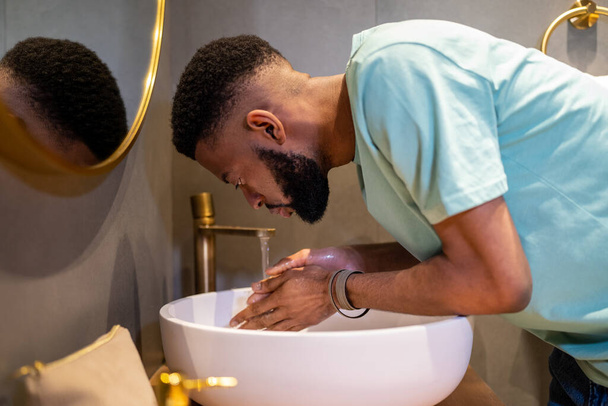 Κουρασμένος νυσταγμένος Αφροαμερικανός που πλένει το πρόσωπο με νερό το πρωί, μαύρος που στέκεται στο μπάνιο και κάνει καθημερινή υγιεινή στο σπίτι πριν πάει στη δουλειά. Καθαρισμός προσώπου για σκούρο δέρμα - Φωτογραφία, εικόνα
