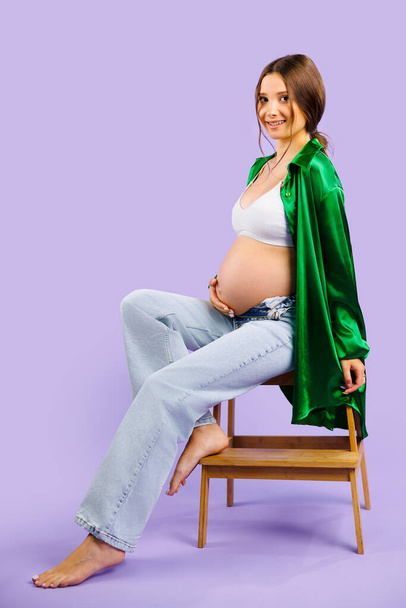 Young beautiful pregnant woman in white stylish body standing, embracing belly smiling looking down over pastel violet background. High quality photo - Foto, Imagen