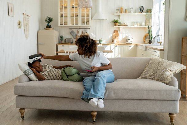 African american boy sleeps in virtual reality glasses on couch at home after long play games mom takes off headset sits near. Tired exhausted dreamy teen child gamer has vr online gaming addiction.  - Foto, Imagen
