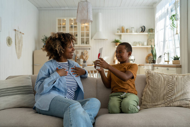 Gelukkige Afro-Amerikaanse familie moeder en zoon vieren samen Moederdag thuis, zittend op de bank in de woonkamer. Kleine jongen kind verrassen moeder met handgemaakte wenskaart. Feestdag - Foto, afbeelding