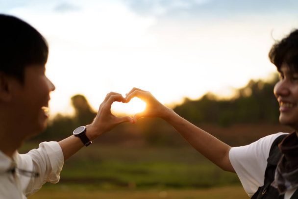 Lovely Asian male LGBTQ couple making a hands in heart shape in a beautiful screen of sunset sky in background. - 写真・画像