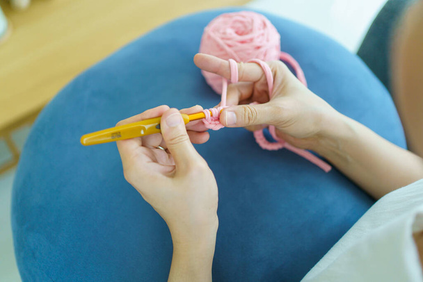 幸せな陽気なアジアの女性は自由時間にリビングルームでかぎ針編みをしています。熟練したアジアの職人の女性は、居心地の良いリビングルームで手編みのかぎ針編みのセーターと帽子を編んで. - 写真・画像