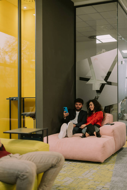 Beautiful colleagues, a couple, lying on pink sofa at modern co working space. They are watching something on the phone and having fun - Φωτογραφία, εικόνα