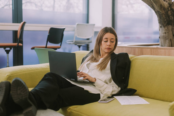 Крупним планом блондинка бізнес-леді друкує і працює на ноутбуці, сидячи на жовтому дивані в робочому просторі
 - Фото, зображення