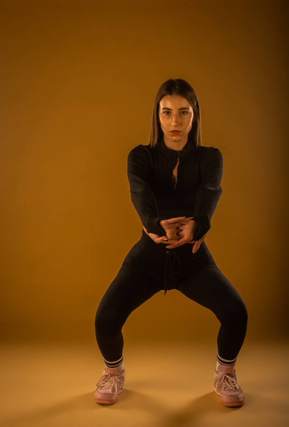 A front view photo of a girl in black sportswear doing squats in studio on a beige background - Fotografie, Obrázek