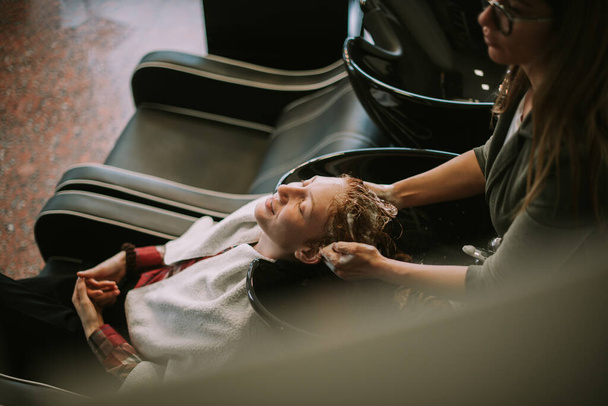 Hermosa chica pelirroja relajándose en la peluquería mientras su peluquera se lava el pelo - Foto, imagen