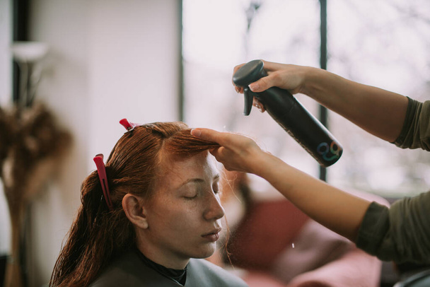 Close up of hairdresser putting hairspray on her red haired client at the hair salon - Photo, Image