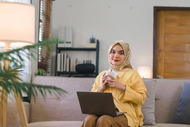 彼女のリビングルームでソファに座っている間、彼女の家のオフィスから働いている黄色のヒジャーブを身に着けている素晴らしいイスラム教徒の千年ビジネス女性 - 写真・画像